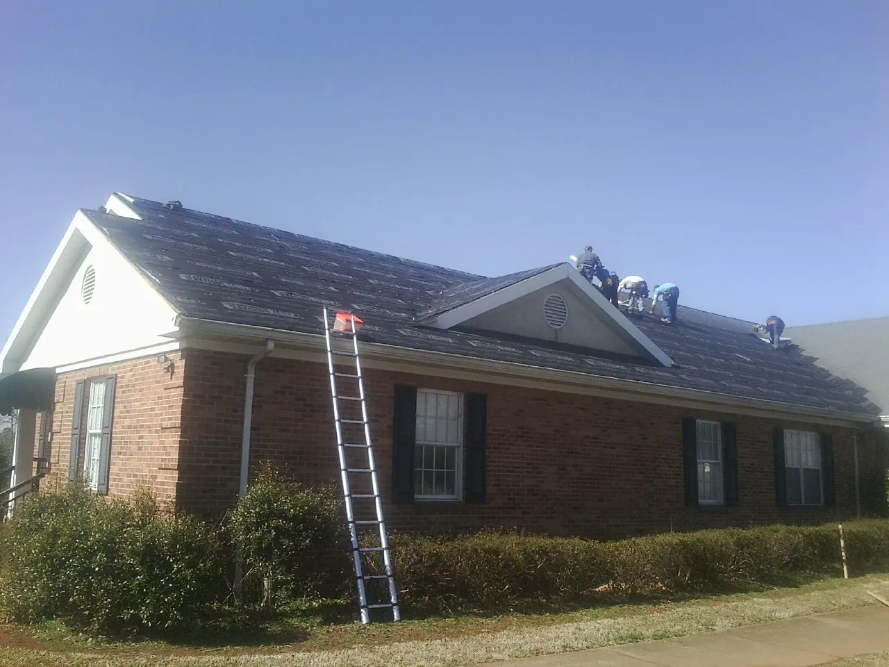 Crew tearing shingles off a brick ranch roof with a ladder set — Emergency Roof Repair work by our crew in Enigma