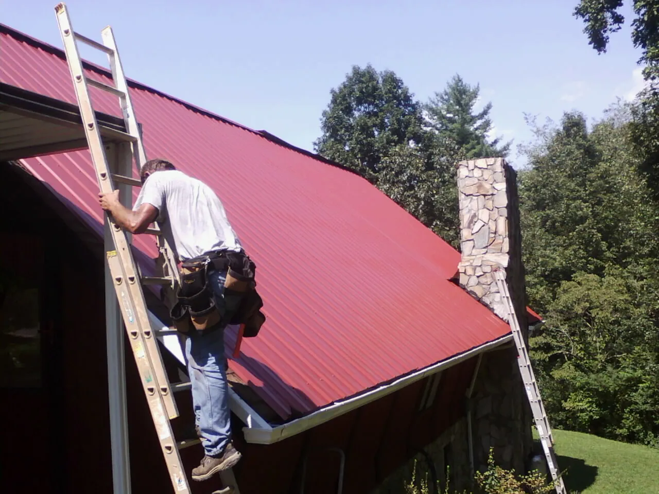 Roofer on a ladder beside a red metal roof and stone chimney — Soffit & Fascia Repair by Roofing Brothers in Enigma